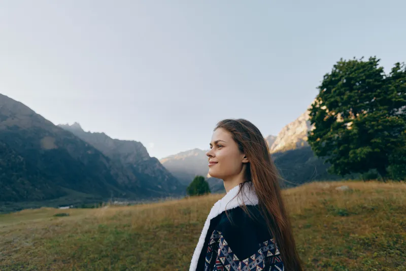 Woman portrait in mountains meadow nature outdoors, young woman in profile smiling with long hair, scenic landscape background with grassy field, lone tree and distant mountain peaks.