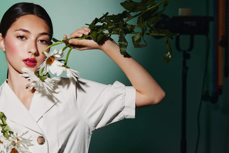 Young woman in a white outfit holds delicate flowers near her head in a fashion photography studio. Creative beauty shot shows elegance and modern style.
