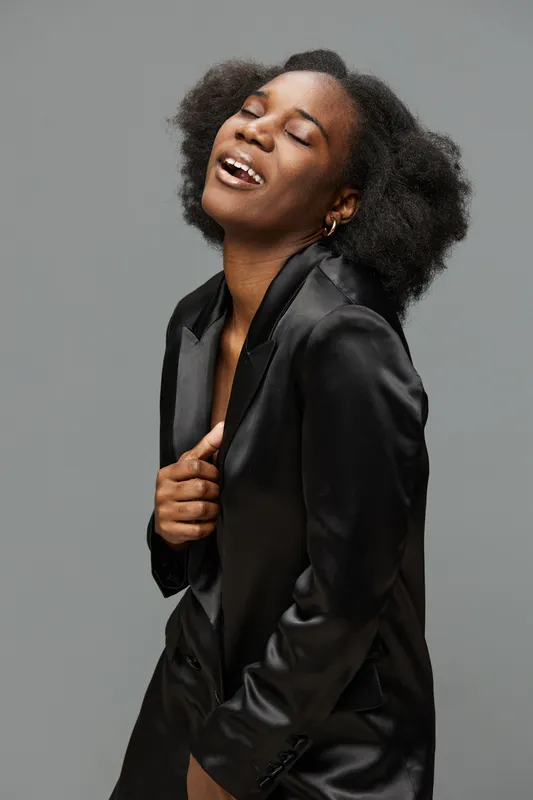 A joyful young Black woman in a stylish black blazer, emoting happiness against a neutral gray background, embodying contemporary fashion and confidence