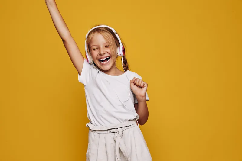 Cheerful girl with blonde hair wearing headphones is dancing and singing with eyes closed on a yellow background. Concept for music, joy, and childhood fun.