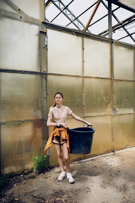 Girl in greenhouse carrying a large bucket for gardening and plants, casual young woman with soil on sneakers and shorts, standing by translucent wall in a summer horticulture portrait.