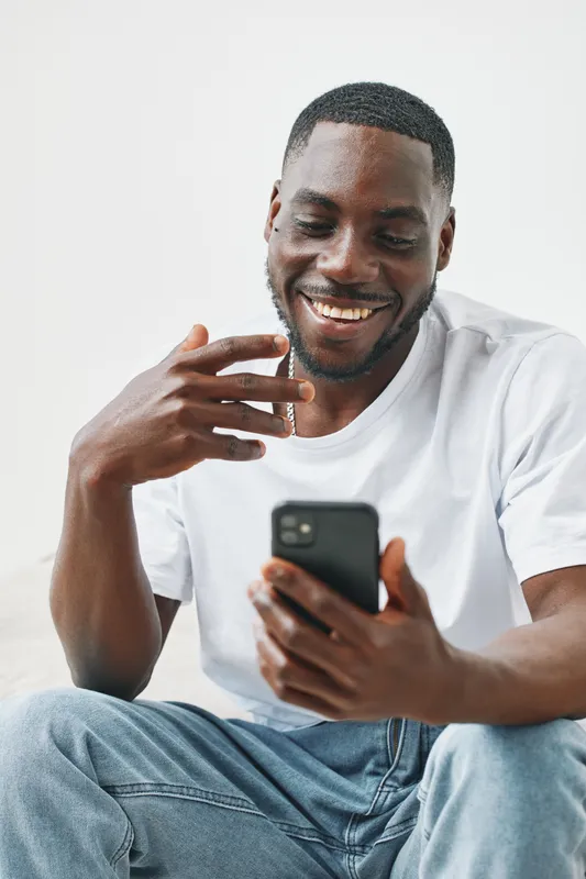 A joyful Black man wearing a white t shirt interacts with his smartphone, showcasing happiness and connection in a bright, minimalistic setting