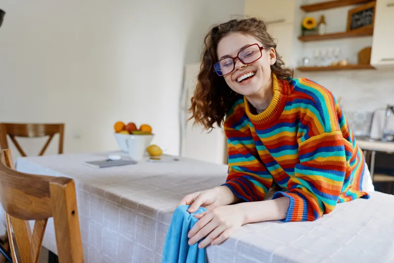 Young woman with curly hair sits at a bright kitchen table wearing a colorful striped sweater and glasses, smiling warmly as she leans forward in a cozy home setting with fruit on the counter and