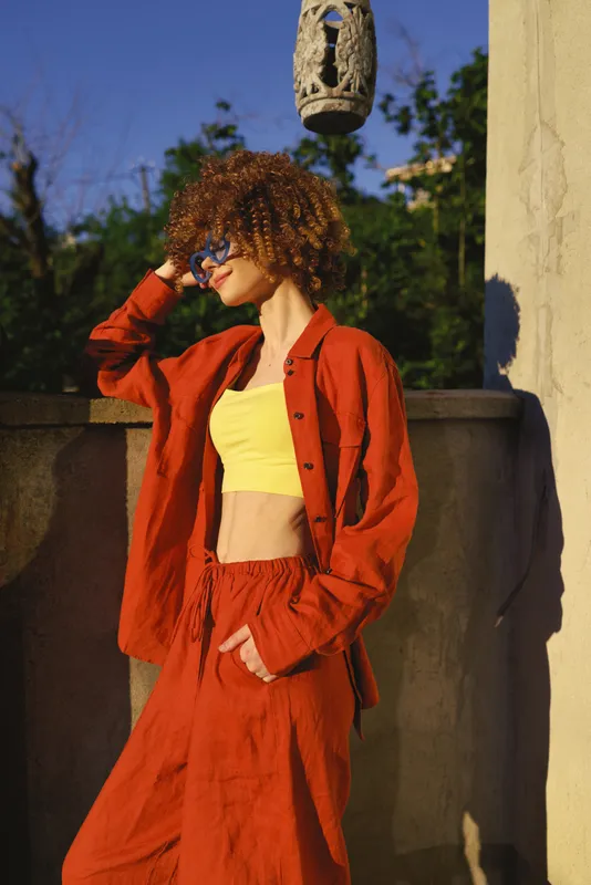 A stylish woman in an orange suit and yellow crop top stands outdoors, shielding her eyes from bright sun. A casual lifestyle moment captured in natural light