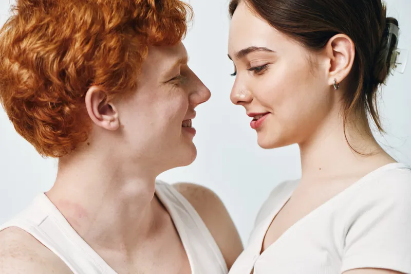Couple with red hair man and brunette woman in white tops looking at each other with affection and smiling on light background. Closeup, love and intimacy concept.