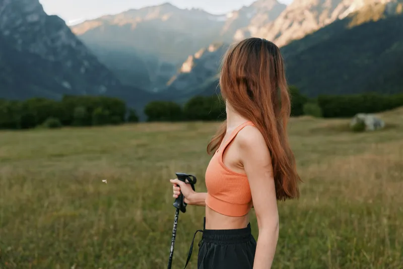 A woman stands in a grassy field with distant mountains, holding a trekking pole. She wears an orange crop top and dark pants, facing sideways with a relaxed, focused expression.