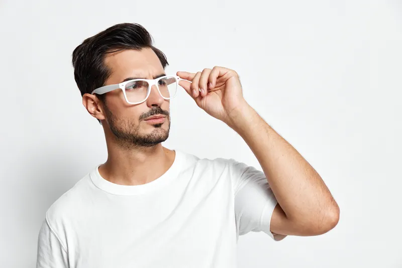 Thoughtful young man wearing white glasses adjusts frames while looking to the side, dressed in a casual white t-shirt, isolated on plain background. People lifestyle concept