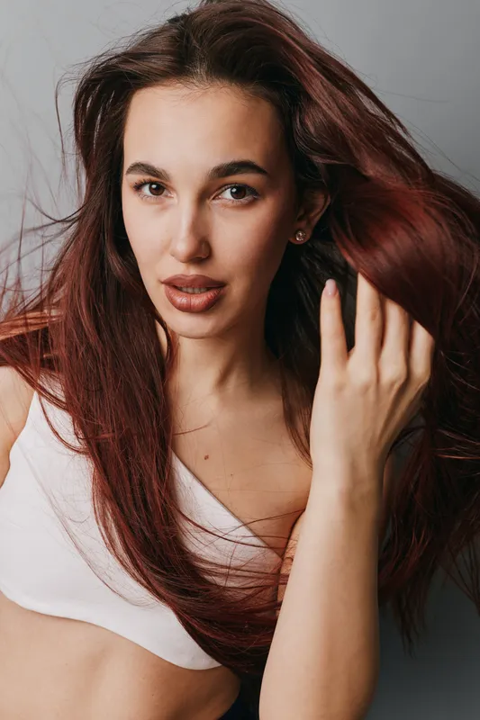 Woman with long hair and natural makeup posing in white top against gray background. Portrait of young female model with expressive eyes and hand in hair for beauty concept.