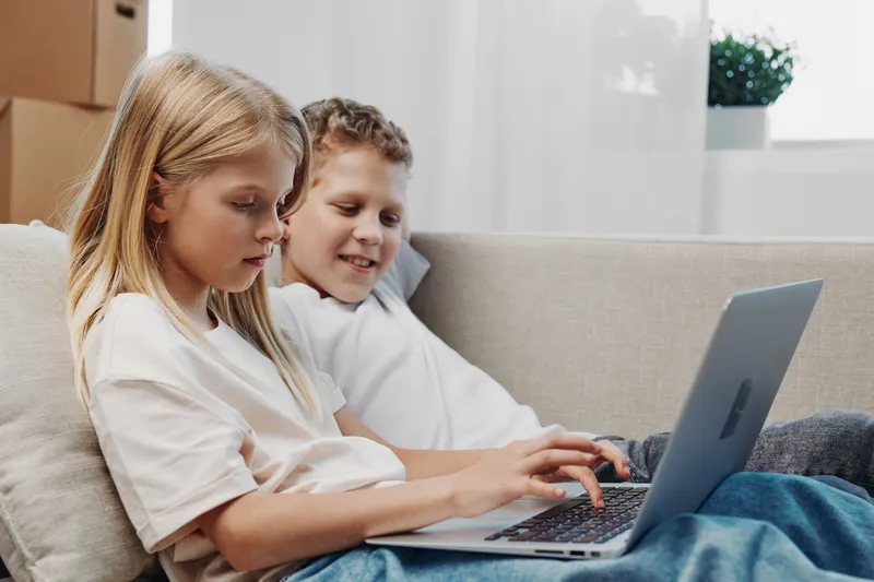 Two children sitting on a sofa, focused on a laptop screen, engaged in learning and technology at home in a bright, cozy living room environment.