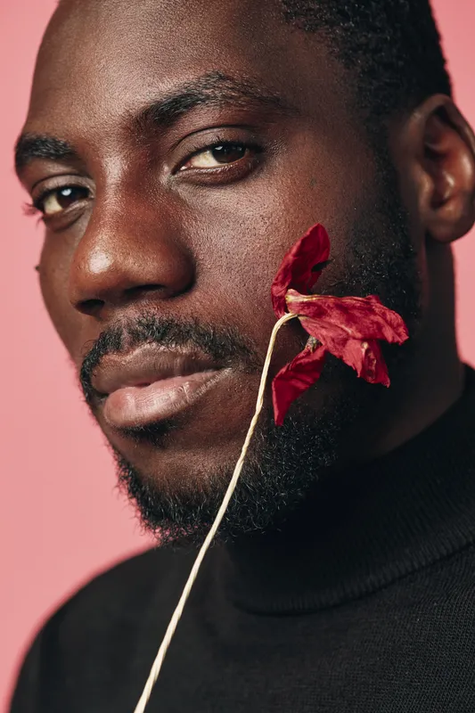 African man with dark skin and beard holding dried red flower near face in creative conceptual visual style with pink background. Closeup portrait with artistic expression and thoughtful mood.