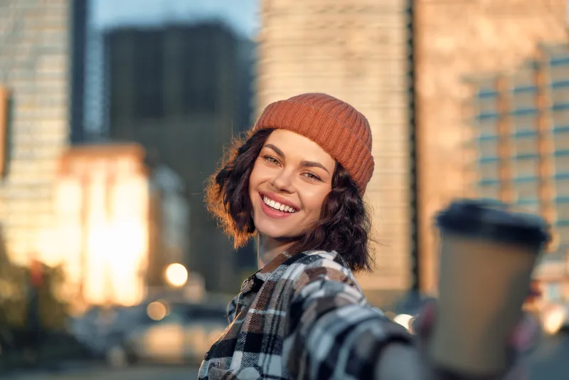 Woman smile in city at golden hour, holding coffee cup toward camera, beanie and plaid jacket, candid lifestyle portrait with authenticity, mindful living and emotional storytelling.