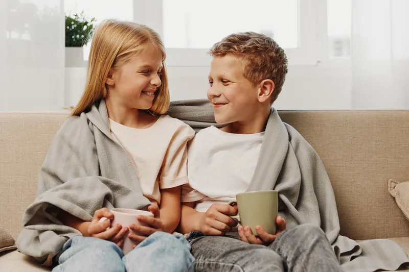 Smiling siblings wrapped in blankets enjoy warm drinks together on a cozy sofa in a bright living room, sharing a happy moment of family bonding.
