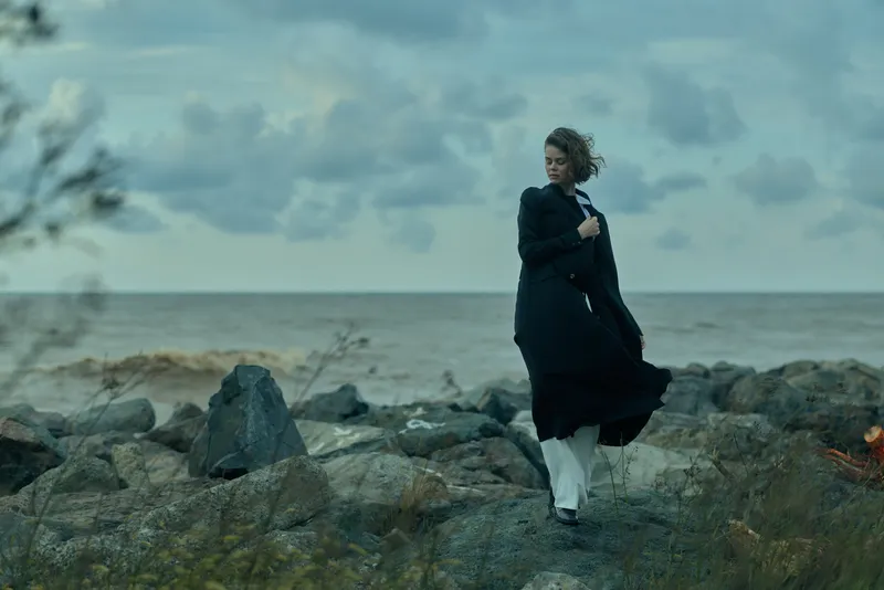 Windswept shoreline with flowing dress and coat along rugged rocks by the ocean, wind and sea create moody outdoor scenery with distant horizon and calm atmosphere