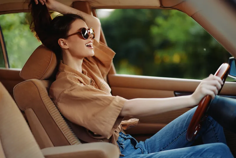 Young woman driving a vintage car, showcasing a carefree and joyful attitude while wearing sunglasses and casual clothing.