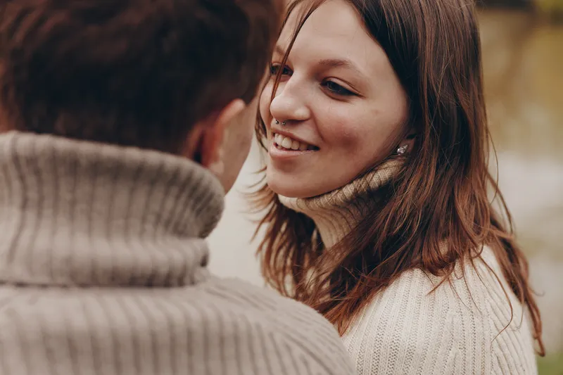 Smiling woman with brunette hair wearing a cozy sweater in a close-up shot, soft light, warm tones capturing a lifestyle moment of affection between partners.