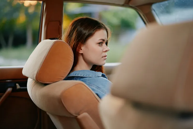 Young woman with brown hair sitting in a vintage car, reflecting on life, wearing a denim jacket, with a soft sunset glow illuminating the background.