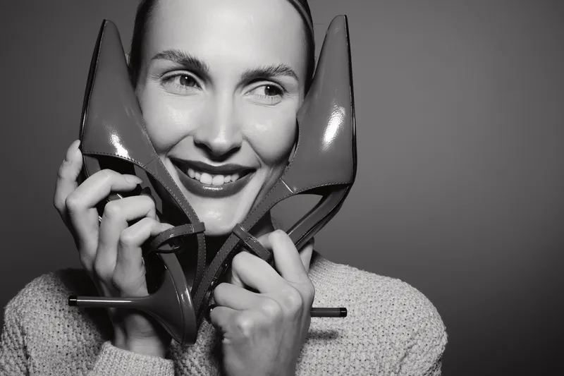 Smiling Caucasian woman with fair skin holds red high heels near face, wearing knitted sweater. Concept of fashion, style, happiness and female beauty in studio lighting.