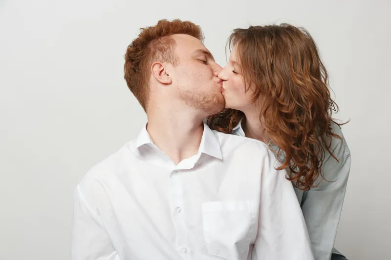 Young couple kissing tenderly on light background. Man in white shirt and woman with curly hair share intimate moment. Concept of love, romance, affection, and relationship.