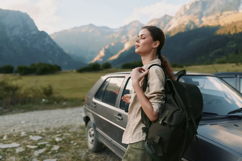 Young woman with a backpack stands beside a parked car in a misty valley, surrounded by mountains and a clear sky, enjoying a quiet moment outdoors.