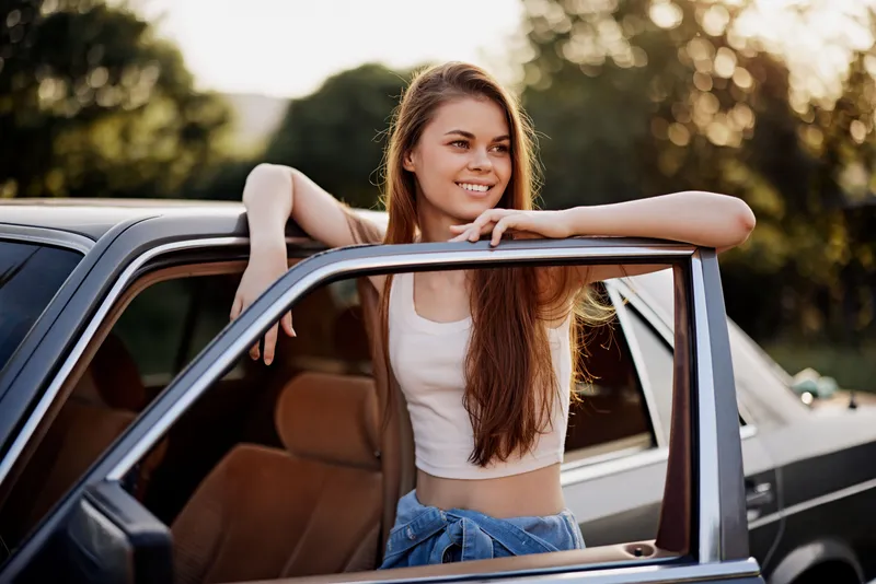 Smiling young woman with long hair leaning out of a vintage car, dressed casually, depicting freedom and joy in a sunny outdoor setting.