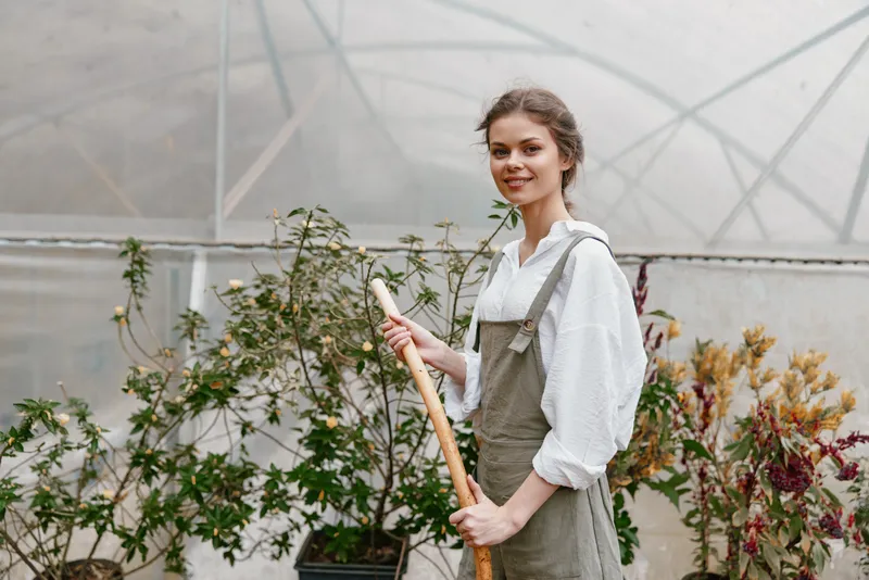 Young woman in a greenhouse wearing an apron, smiling confidently while gardening Her casual outfit and greenery reflect a vibrant spring atmosphere