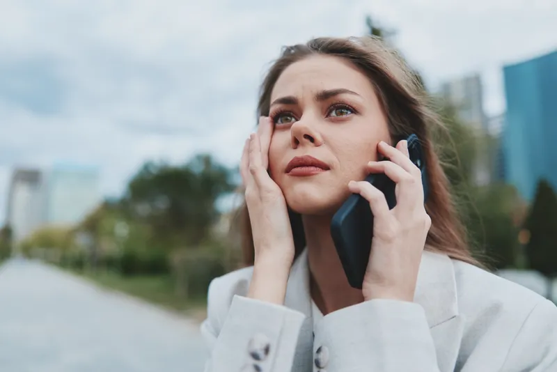 Woman stressed on phone call outdoors, young Caucasian woman in business attire with worried expression against urban background, financial crisis concept