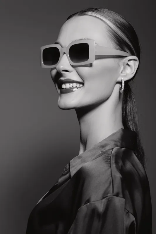 Smiling young white woman with sunglasses and sleek hair posing in studio. Portrait of confident female model with fair skin and elegant style in black and white.