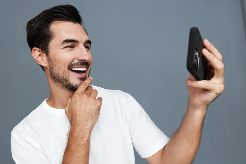 Smiling man with beard wearing white t-shirt looking at smartphone, enjoying video call, isolated on solid grey background. People lifestyle concept.