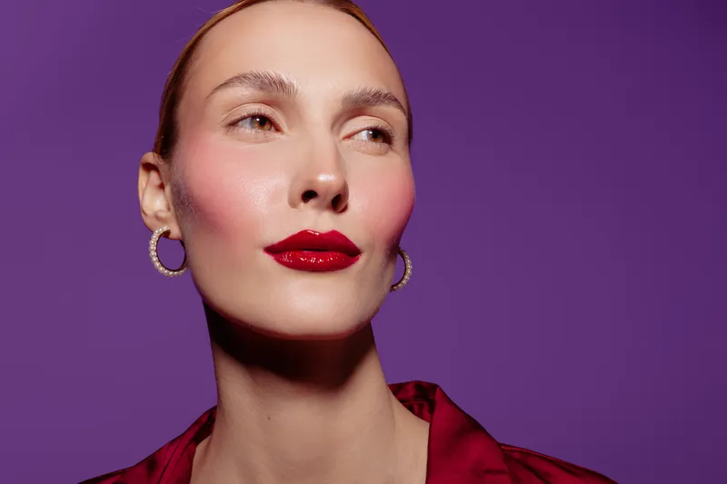 Close-up portrait of confident woman with red lipstick and hoop earrings on purple background. Concept of beauty, makeup, fashion, and elegance with studio lighting.