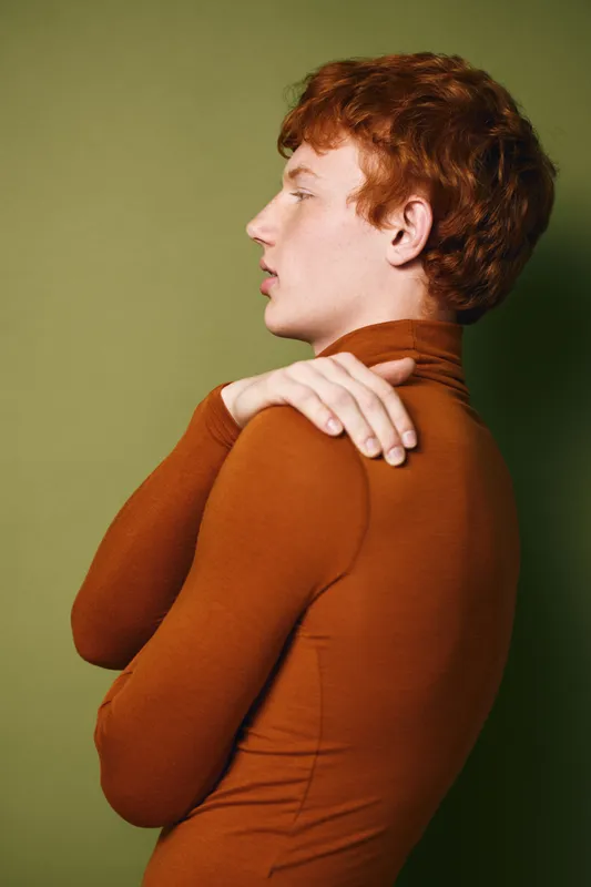 Young man with red hair wearing brown turtleneck standing in profile against green background. Portrait of thoughtful person with hand on shoulder in casual clothing and natural light.