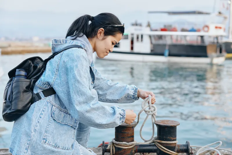 Traveler in a denim jacket ties a rope to a bollard at the harbor, with a backpack, preparing lines for docking, maritime activity by the water and urban seaside atmosphere