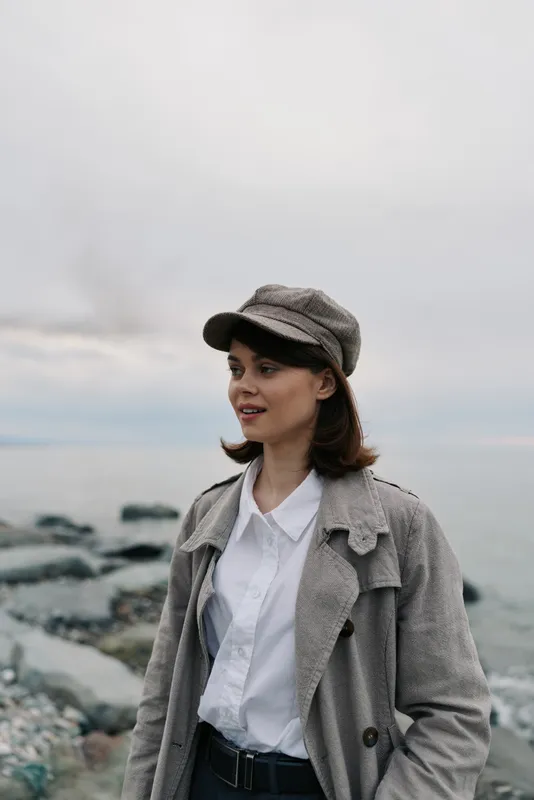 Young woman wearing gray coat and cap stands on rocky beach near calm sea. Overcast sky creates soft light and peaceful atmosphere. Concept of autumn fashion and outdoor portrait.