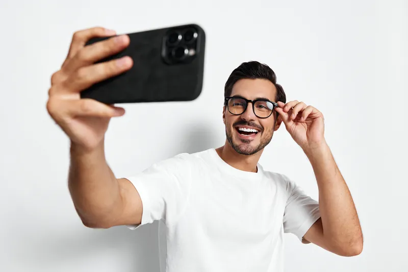 Happy smiling young man taking selfie with smartphone, wearing glasses and casual white t-shirt, capturing joyful moments against plain light background.