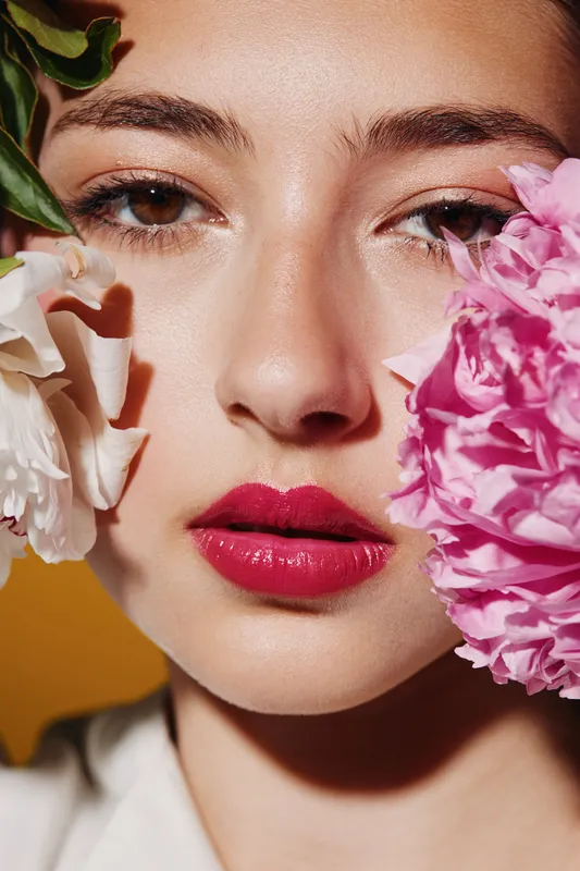 Close-up of a young woman with glossy dark pink lips and natural makeup, framed by pink and white peonies. Concept for beauty, fashion, creative skincare, and fresh floral style.