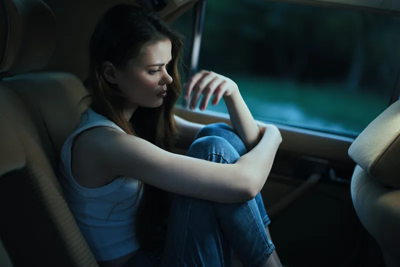 A contemplative young woman sits curled up in a car, reflecting on her emotions as the evening light softly enters through the window, dressed casually in a tank top and jeans.