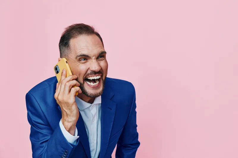 A professional man in a blue suit on a pink background, speaking on a smartphone with a joyful, confident expression that conveys energy and business presence.