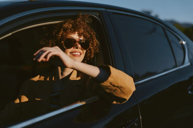 Smiling woman with curly hair leaning from a car window, sunglasses on, enjoying warm sunlight. Casual lifestyle moment with soft retro film color and relaxed mood.