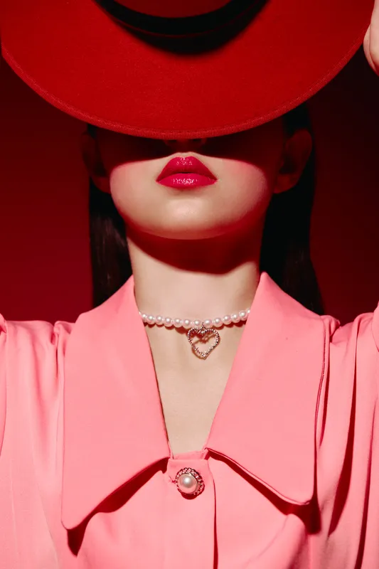 Fashionable woman with shiny red lips hides eyes under hat, wearing pink blouse with pearl pendant necklace. Studio beauty shot with creative lighting on red background, concept fashion.