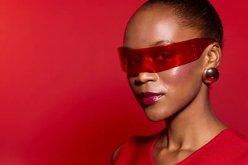 Close-up portrait of confident African woman with dark skin wearing red futuristic glasses and earrings against red background. Concept for fashion, style, and modern beauty.
