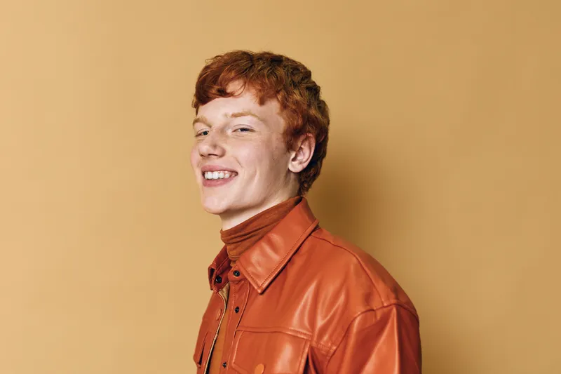 Young man with red hair wearing brown leather jacket smiling and looking at camera against beige background. Casual style, confident pose with natural light and warm tones.