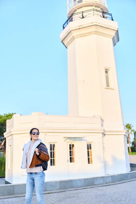 Woman in sunglasses and scarf beside a white lighthouse, outdoor travel scene with casual attire, bag on shoulder, daylight sky and coastal beacon in view