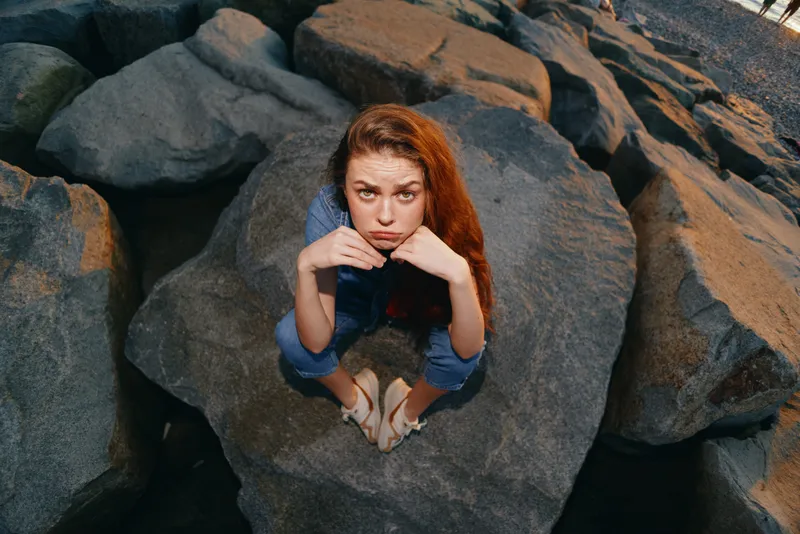lifestyle woman in modern boho-western denim street style sitting on rocks with film color effect and natural red hair in outdoor urban setting