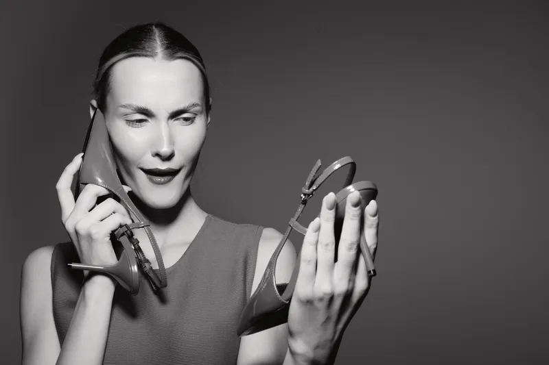 Confused white woman with light skin and slicked hair holds high heel shoe near face, looking puzzled in studio with plain background. Concept for fashion doubts and style choices.
