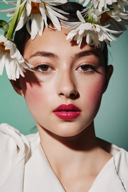 Young woman with vibrant pink lips and natural glow wears daisy flower crown and white blouse. Close-up portrait with soft lighting on solid green background. Concept of beauty and fashion creativity.