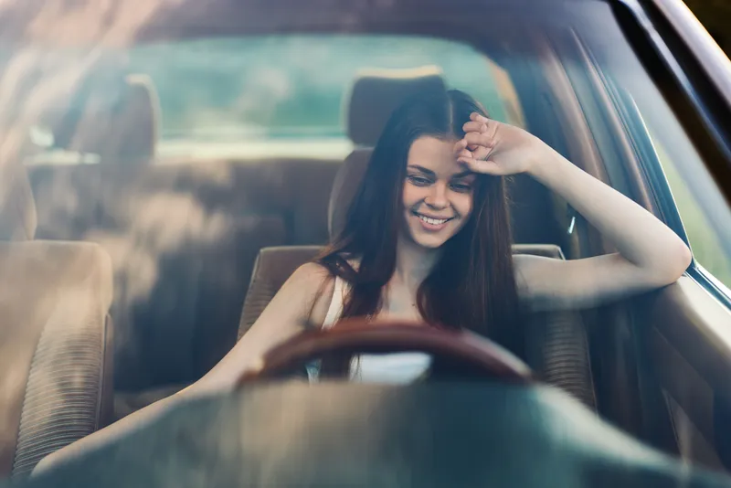 Young woman smiling joyfully while sitting in a car, showcasing carefree emotions and a relaxed atmosphere with sunlight reflecting on her face.