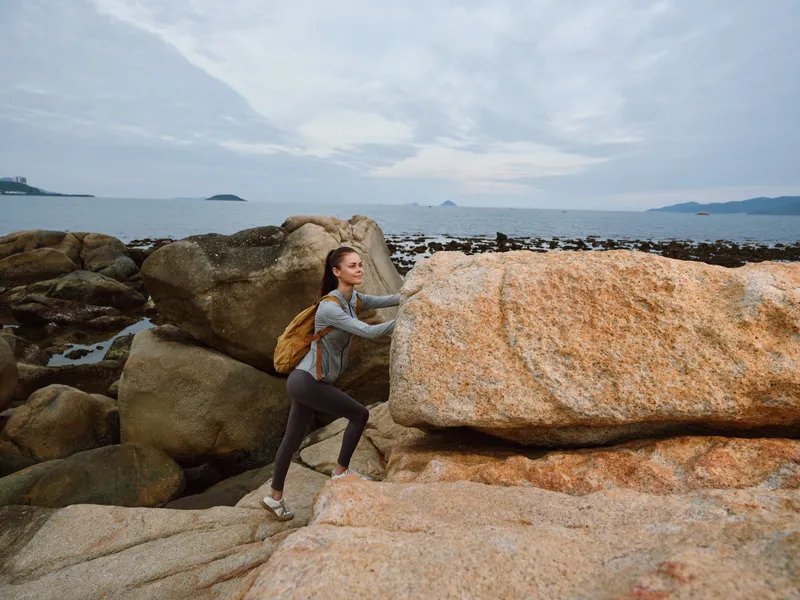 Young woman climbing rocky coastline, showcasing determination and adventure with a scenic sea background, perfect for outdoor and fitness themes