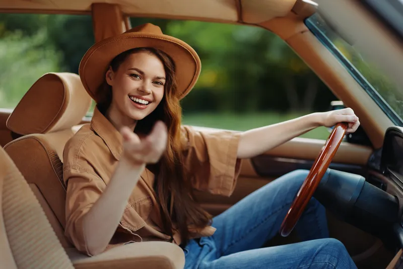Young woman smiling in a vintage car, wearing a hat and casual outfit, surrounded by a green natural background, capturing a sense of freedom and adventure.