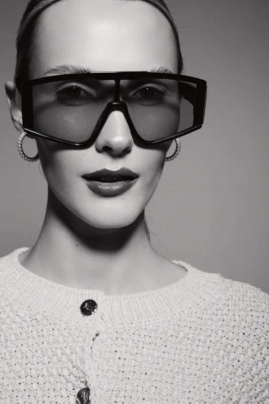 Fashionable white woman with pale skin wearing large sunglasses and hoop earrings poses in textured sweater. Studio portrait with modern style and confident expression.