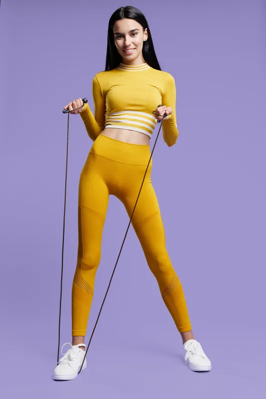 Woman sport fitness in yellow activewear holding a jump rope, posing against a purple background, showing energy, health, and athletic lifestyle in a studio setting.