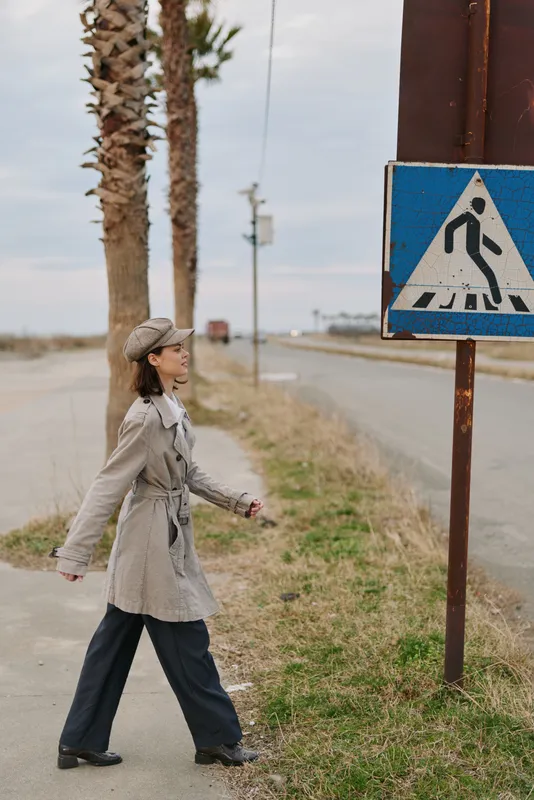 A young woman in a beige trench coat and beret walks along a roadside near palm trees and a pedestrian crossing sign on a cloudy day. Urban fashion and outdoor concept.